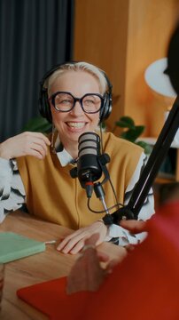 Smiling female podcast host wearing headphones and glasses recording show in studio, speaking into microphone and chatting with guest. Vertical format video