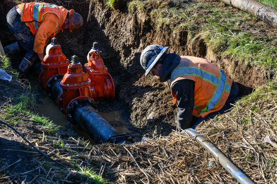 Workman installing a wedge gate valve in municipal water supply distribution pipeline, working in dirt trench on a sunny winter day with water spraying up, new development infrastructure construction
