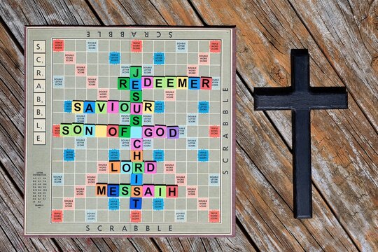 Scrabble game board with names and titles of Jesus on the board. And a dark black wooden cross on the right of it.