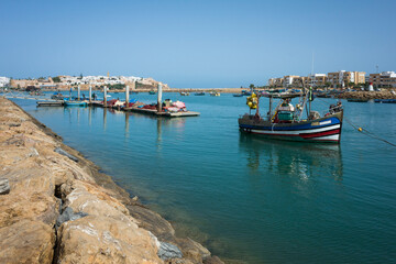 Obraz premium Fishing boat on the river between Rabat and Salé, Morocco. Calm water, docks, and city buildings on both riverbanks under clear blue sky. Peaceful coastal scene with traditional vessel
