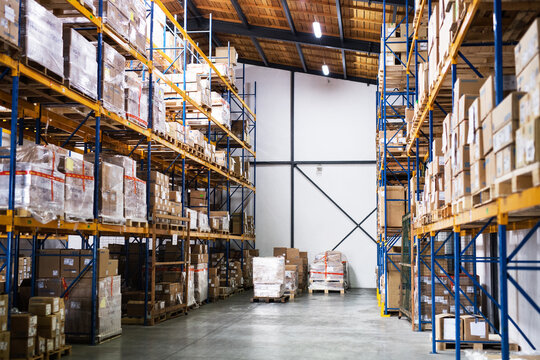 Large warehouse interior with tall pallet racks stacked with cardboard boxes and goods, illustrating organized storage, inventory management and modern logistics operations

