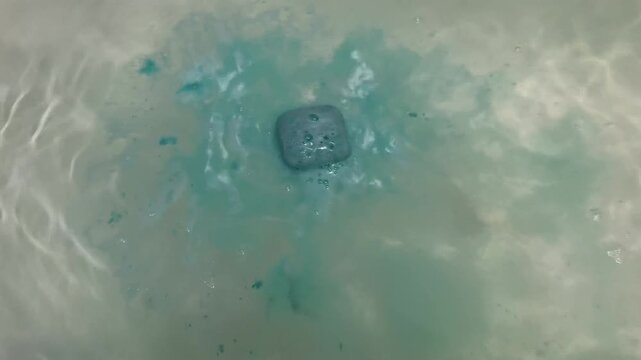 Female hand dropping a blue bath bomb into a filled bathtub. The bath bomb fizzes and bubbles in the water. Relaxing self-care moment.