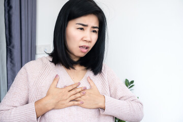Asian woman holding chest in pain, suffering from heart attack or acid reflux symptoms at home. Young female feeling chest tightness, heartburn, or breast pain. Healthcare and emergency concept