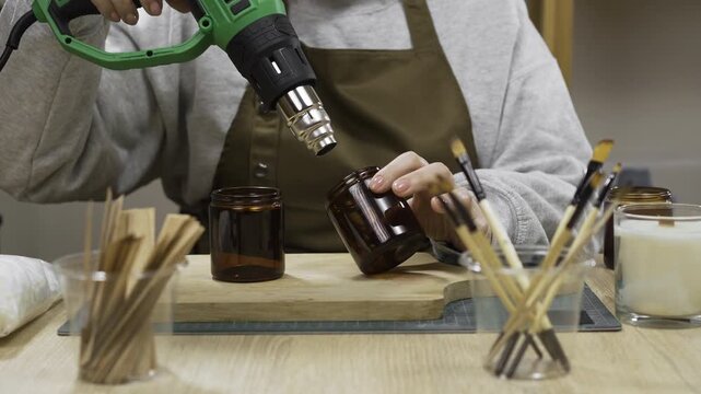 female hands heating glass jars while making homemade wax candles in a workshop using a heat gun.