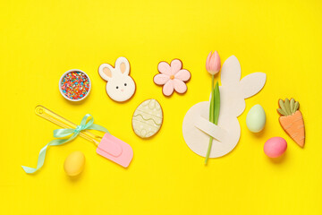 Composition with tasty Easter cookies, spatula and bowl with sprinkles on yellow background