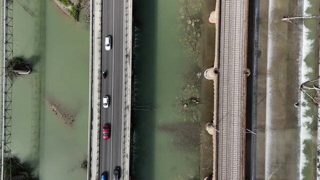SS10 highway bridge crossing the Trebbia River near Piacenza, Emilia-Romagna, with cars in motion above green water beside the parallel brick railway viaduct and riprap banks, top down shot