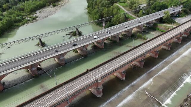 SS10 highway bridge crossing the Trebbia River near Piacenza, Emilia-Romagna, with cars in motion above green water beside the parallel brick railway viaduct and riprap banks