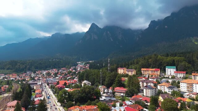 Drone shot advancing above Busteni town, showing residential buildings with the Bucegi Mountains rising in the background under a cloudy sky.