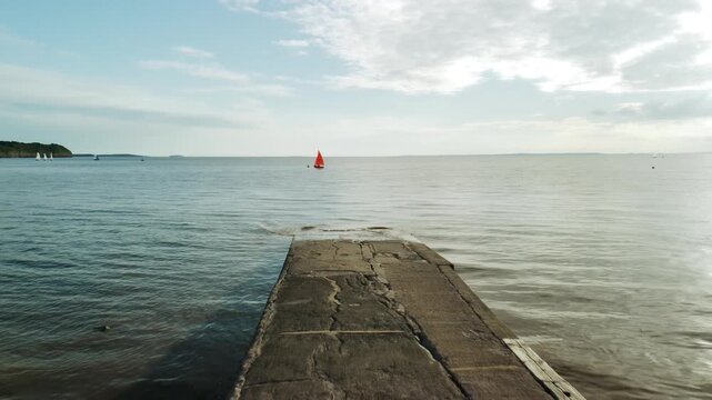 Drone shot of a concrete slipway leading into a calm sea, with sailboats on the horizon and a bright red sail under a soft, clouded sky &mdash; peaceful coastal atmosphere. Shot tracks forward towards boat