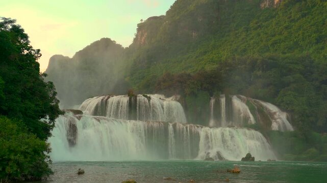 A wide shot of the multi-tiered Ban Gioc Waterfall flowing into a turquoise river, framed by dense jungle and limestone cliffs during dusk.