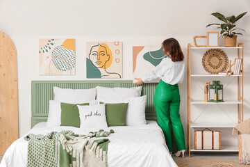 Young woman hanging painting on light wall in bedroom, back view