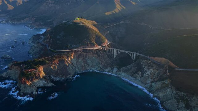 Aerial view of big sur coastline bridge, golden hour light bathing Bixby Creek Bridge and rugged cliffs, Pacific waves crashing into sheltered cove, mist drifting over coastal road, sweeping