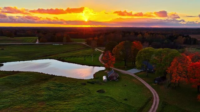 Aerial: fire sky sunset over a forest with a small lake and a house, pull out drone shot