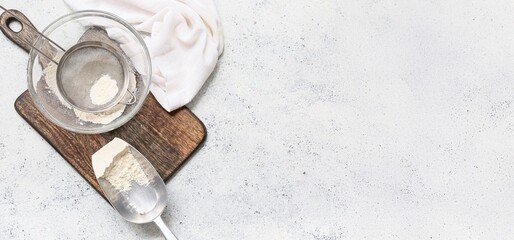 Bowl with flour, metal sieve, scoop and wooden board on light grunge background with space for text