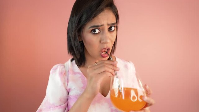 Close up portrait of surprised pretty young Indian woman lady holding glass sipping drinking orange juice staring at camera with amazed face expression, isolated over pink studio background.
