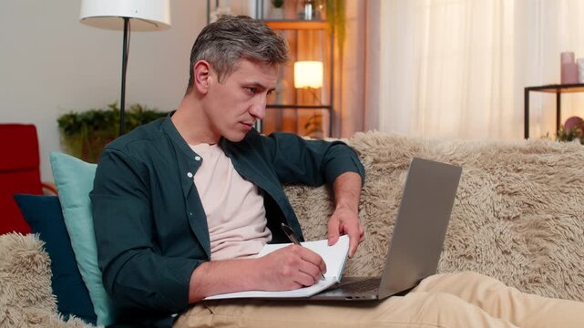 Young man at home calmly writes in notebook while reading from laptop with focused face. Caucasian guy works attentively and efficiently on sofa, staying concentrated and serious about task at hand.