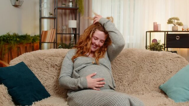 Pregnant young woman sits on home sofa, gently touching her belly, laughing with bright smile, embracing positive mood. Girl feels happiness and connection, finding joy in small moments of motherhood