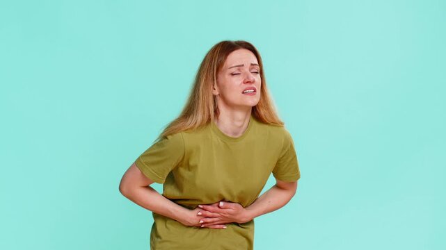 Middle-aged woman holds stomach with visible pain, expressing discomfort from cramps, menstrual pain, or bad food reaction. Girl isolated on blue background shows signs of stomach ache or poisoning.