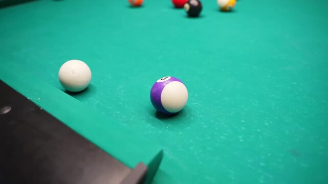 Cue ball and purple stripe sit near the pocket on a green pool table, with other balls blurred in the background under cozy bar lighting and shallow depth of field.