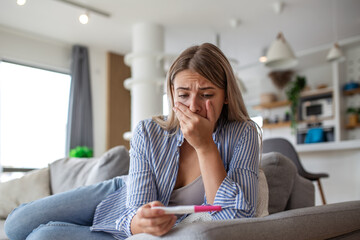 Worried woman looking at pregnancy test. Unwanted pregnancy or fertility problem concept. Sad girl holding positive test sitting on sofa at home. Healthcare and reproductive medicine concept.