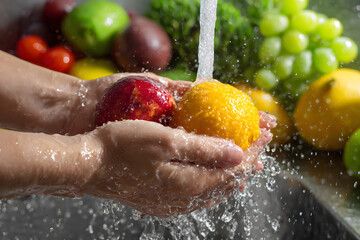 Washing fresh fruits in hands under running water in the kitchen sink with colorful produce in the background