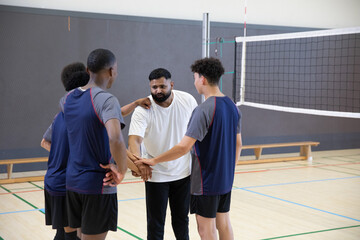Diverse teenage males in navy jerseys surrounding Indian coach placing hand into huddle at gym net © wavebreak3