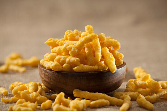 Crispy cheese curls corn snacks in a wooden bowl, extruded cheese puffs scattered on a rustic burlap surface.