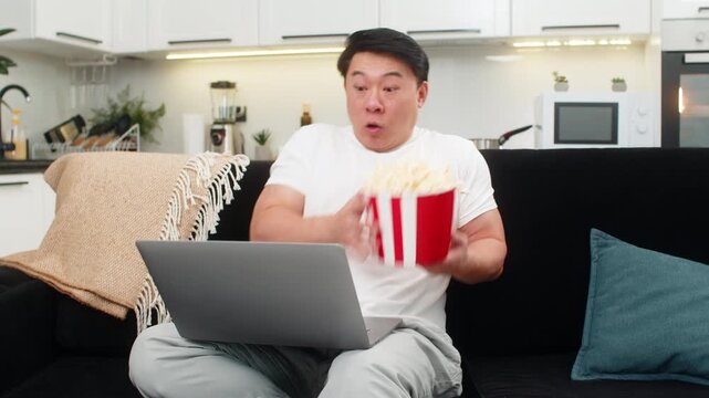 Asian man at home watches scary thriller on laptop jumps at scare spills popcorn then breathes and slows pulse. Chinese guy on sofa keeps watching curious holds backet firm and waits for big twist