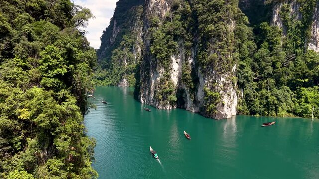 In Khao Sok National Park, a longtail boat glides through tranquil emerald waters surrounded by towering limestone cliffs, revealing the splendor of Thailand natural beauty under a clear blue sky.