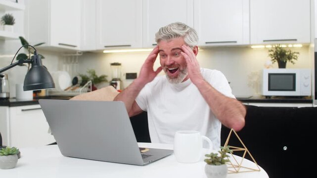 Middle-aged man at home uses laptop sees discount and promo code success smiles. Mature guy sitting at table on sofa cheers small win celebrates online game result lottery casino jackpot news smiles
