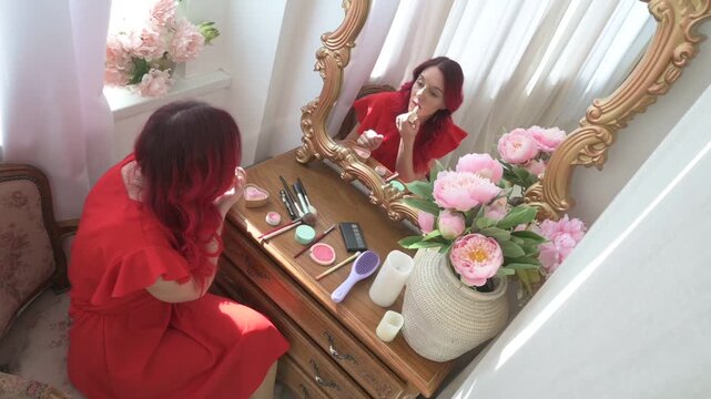 Beautiful woman with red hair applying makeup at an ornate vintage vanity table