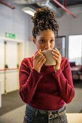 African American woman sipping from beige mug in office breakroom wearing red sweater and jeans