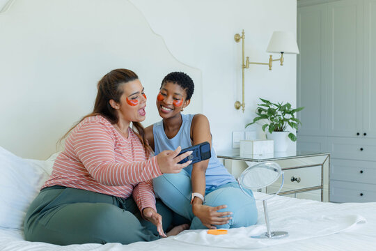 African American female friends sitting on bed wearing orange under-eye patches and checking phone