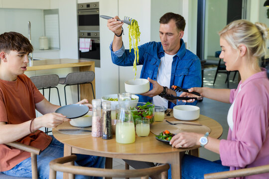 Family with parents and teenage son serving at round wooden table, man lifting pasta with spoon