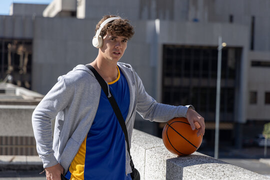 Teen standing on parking roof with headset and jersey, leaning on concrete ledge holding basketball