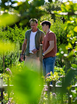 Father and teenage son standing in garden inspecting raised beds, holding paper, wearing smartwatch