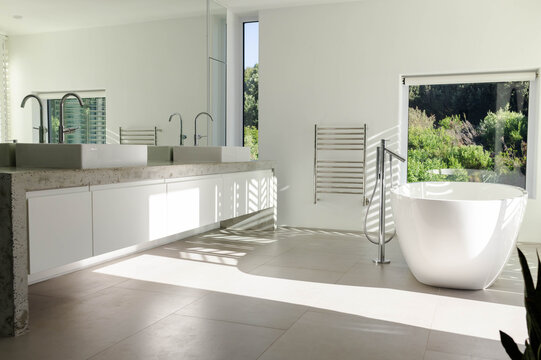 Freestanding white oval bathtub and floating vanity are gleaming under sunlight with garden view