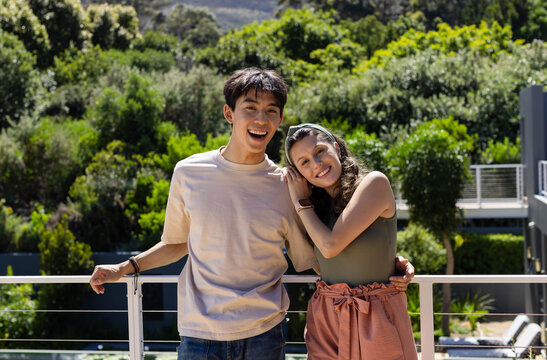 Diverse couple standing on balcony, leaning close against metal cable railing, smiling