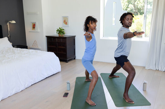 African American couple lunging on mats in bedroom in sportswear facing window with water bottles