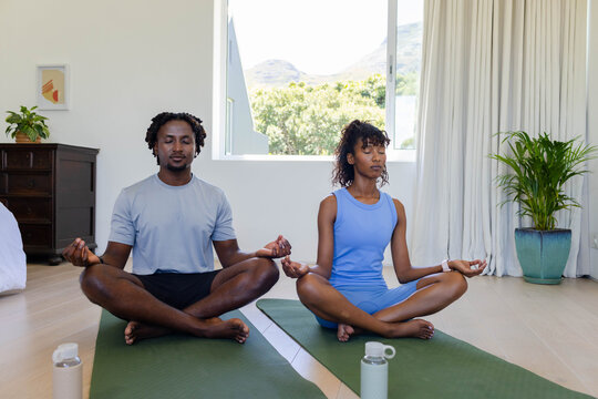 African American couple meditating in sportswear on green yoga mats with water bottles by window