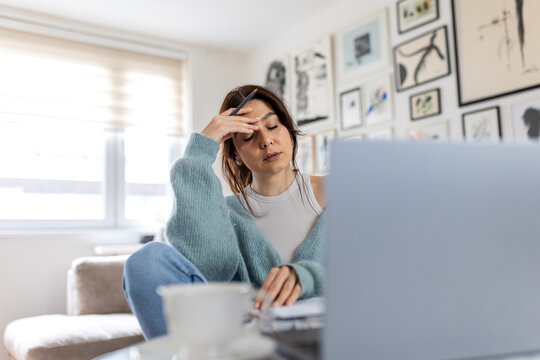 Stressed businesswoman with eyes closed touching forehead while working on laptop. Tired female entrepreneur suffering from headache or burnout in home office. Overworked and fatigue concept.