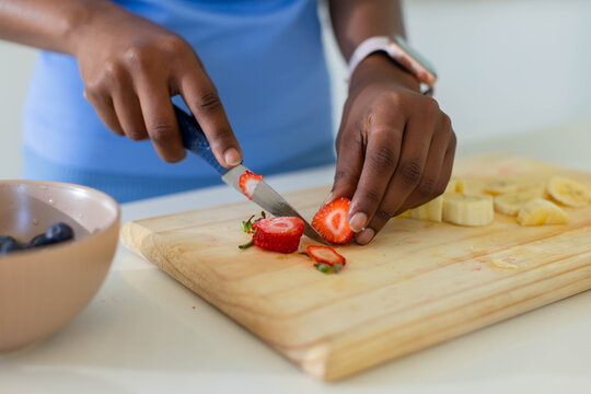 Hands are slicing ripe strawberry with small paring knife on wooden cutting board, blueberries left