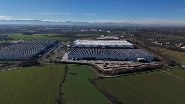 Drone establishing view of a large logistics and industrial warehouse in San Nazzaro, Province of Piacenza, with a full rooftop solar panel system, beside SS10 and surrounded by Po Valley farmland.
