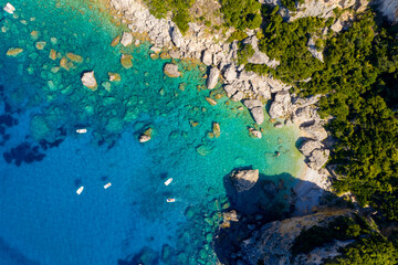 Paradise found: an aerial top-down drone perspective of a secret rocky beach and pristine turquoise sea in Corfu, Greece. Small boats float on the crystal clear Ionian water