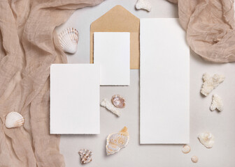 Fototapeta premium Cards and envelopes on beige stone with seashells top view, mockup. Coastal wedding stationery set