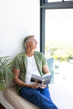 Mature male sitting on soft window bench holding paperback near palm plant and window