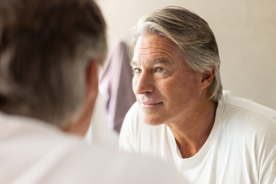 Senior man standing at bathroom mirror inspecting face with towel nearby while wearing white tee