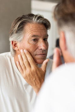 Mature male examining face in bathroom mirror, touching cheek and wearing plain white T-shirt