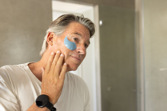 Senior man applying blue under-eye gel patch while checking skin in bathroom mirror