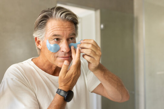 Mature man applying blue hydrogel under-eye patches at bathroom mirror, wearing white t-shirt